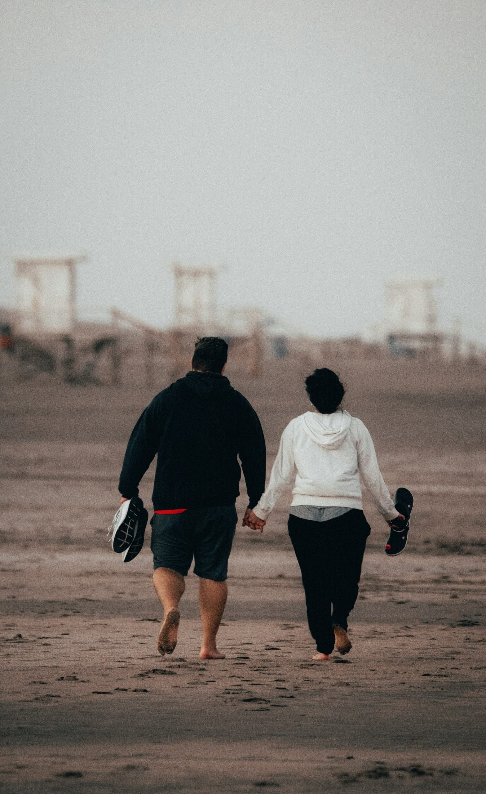 Man and woman holding hands walking on the beach with sneakers and a yoga mat in hand.